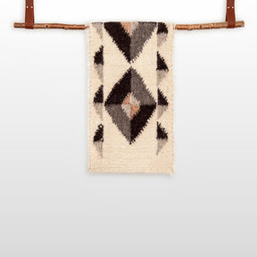 The Iskara (Spark) Wool Bed / Couch Runner, featuring black and brown geometric patterns, is handcrafted and displayed hanging on a wooden rod against a white background.