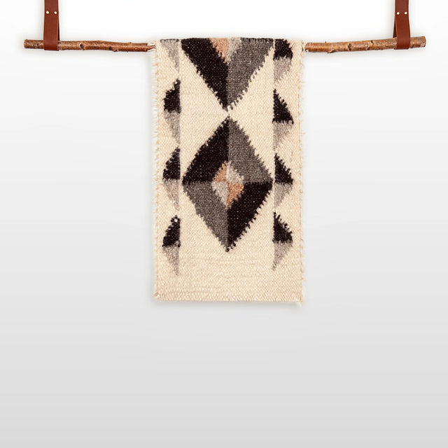 The Iskara (Spark) Wool Bed / Couch Runner, featuring geometric diamond patterns, hangs from a wooden branch with leather straps against a plain background.