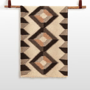 The Svitlo (Bright) Wool Wall Hanging features a beige base with geometric diamond patterns in brown, black, and tan, displayed on a wooden rod—a stunning piece of handcrafted woven wall art.