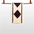 The Korysno (Helpful) Wool Bed / Couch Runner is crafted from organic Carpathian wool, featuring dark diamond patterns on cream fabric and draped over a wooden rod—ideal for sustainable home decor.