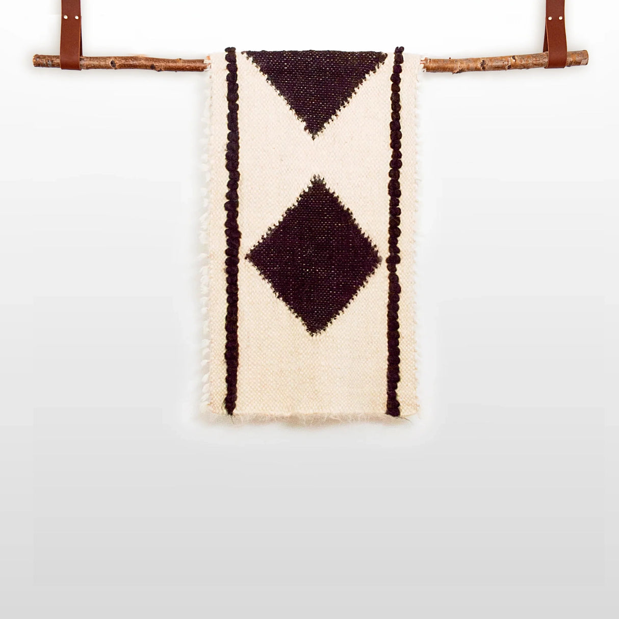 The Korysno (Helpful) Wool Bed / Couch Runner is crafted from organic Carpathian wool, featuring dark diamond patterns on cream fabric and draped over a wooden rod—ideal for sustainable home decor.
