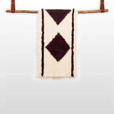 The Korysno (Helpful) Wool Bed / Couch Runner is crafted from organic Carpathian wool, featuring dark diamond patterns on cream fabric and draped over a wooden rod—ideal for sustainable home decor.