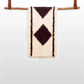 The Korysno (Helpful) Wool Bed / Couch Runner is crafted from organic Carpathian wool, featuring dark diamond patterns on cream fabric and draped over a wooden rod—ideal for sustainable home decor.
