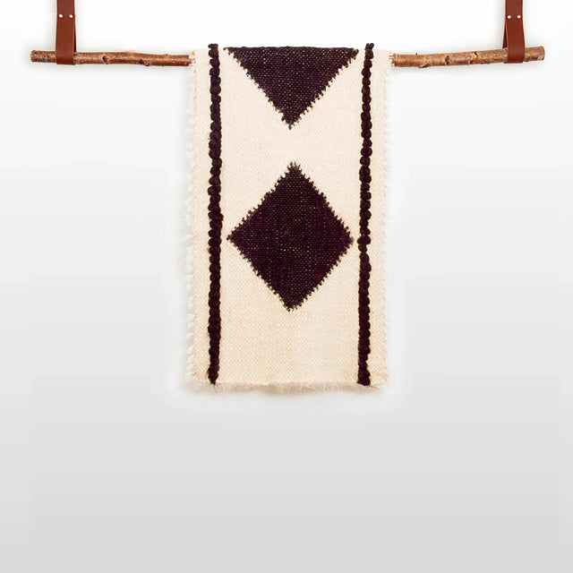 The Korysno (Helpful) Wool Bed / Couch Runner is crafted from organic Carpathian wool, featuring dark diamond patterns on cream fabric and draped over a wooden rod—ideal for sustainable home decor.
