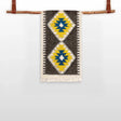 The Mriya (Dream) Wool Bed / Couch Runner, featuring yellow, blue, and white diamond patterns and Ukrainian craftsmanship, hangs from a wooden rod with leather straps.
