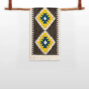 The Mriya (Dream) Wool Bed / Couch Runner, featuring yellow, blue, and white diamond patterns and Ukrainian craftsmanship, hangs from a wooden rod with leather straps.