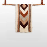 The Tkach (Weaver) Wool Bed / Couch Runner features brown and tan chevron patterns in undyed wool, displayed on a wooden rod with leather straps.