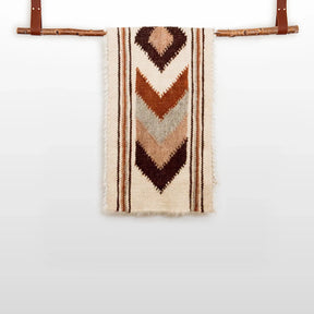 The Tkach (Weaver) Wool Bed / Couch Runner features brown and tan chevron patterns in undyed wool, displayed on a wooden rod with leather straps.