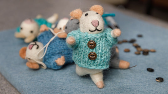 A felt mouse toy in a blue sweater stands upright, with other felt mice and buttons scattered in the background.