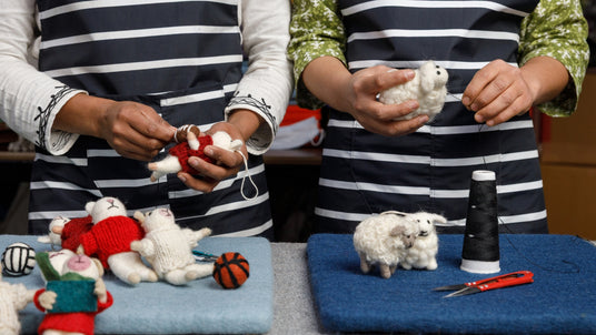 Two people in aprons hand-sew small stuffed animals on blue mats with thread, scissors, and felt.