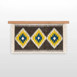 The Mriya (Dream) Wool Wall Hanging, handwoven from Carpathian wool, showcases yellow, blue, and white diamond patterns on a wooden rod—an ideal sustainable home decor piece.