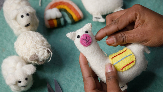 Hands sewing a felt llama among other felt animals and a rainbow on a green surface.