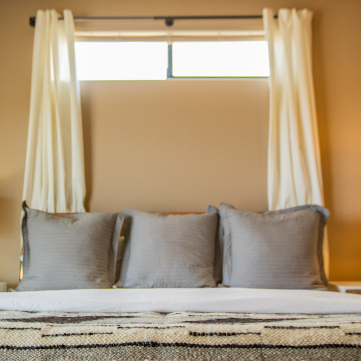 A neatly made bed with three gray pillows, white sheets, and the Tak (Yes) Wool Statement Bed Blanket adds a cozy touch, while cream curtains frame a small window above.