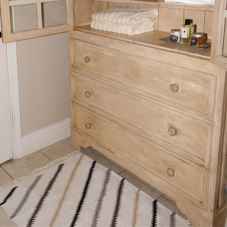 A beige wooden dresser with folded towels, toiletries, and a Promin (Beam) Wool Rug / Wall Hanging beside a patterned rug on tiled flooring.