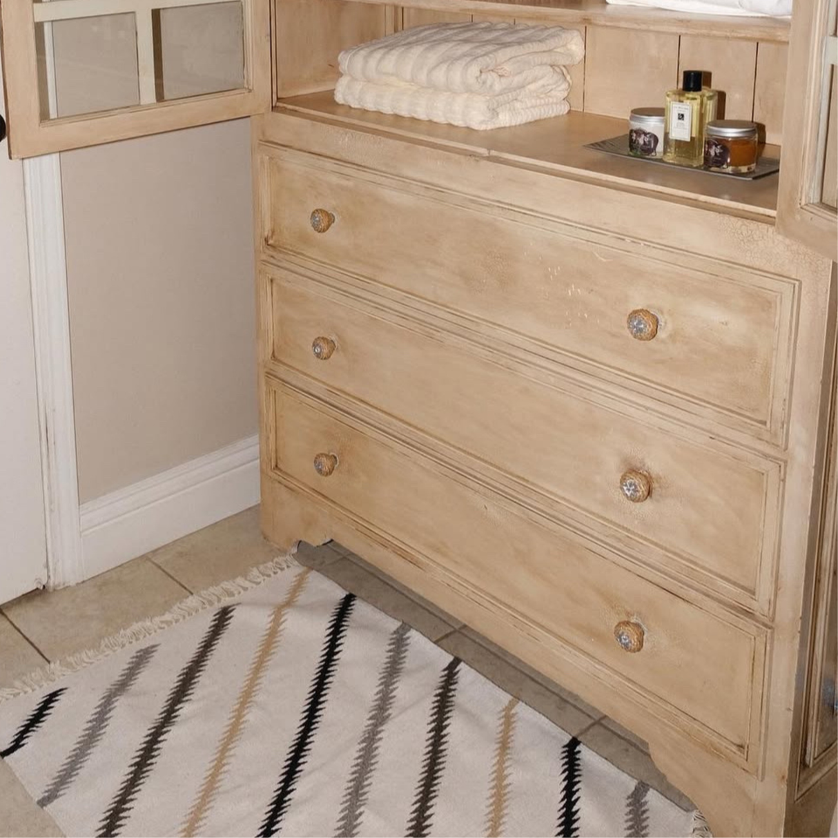 Light wood dresser with three drawers, towels and toiletries on top, and the Promin (Beam) Wool Rug / Wall Hanging—handwoven and inspired by Carpathian mountain sheep—in front.
