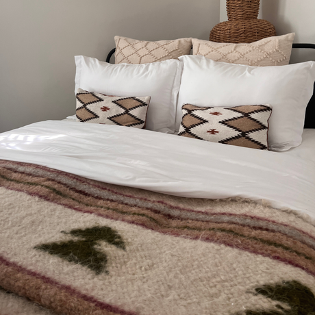 A neatly made bed with white pillows, patterned cushions, and a cozy Sosna (Pine) Wool Statement Bed Blanket featuring tree designs.