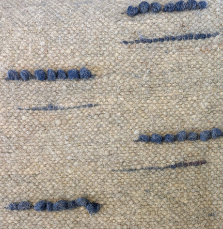 Close-up of the Horoshy (Kind) Wool Statement Bed Blanket: textured beige fabric, handwoven from organic cruelty-free wool, features small uneven rows of blue yarn stitched throughout.