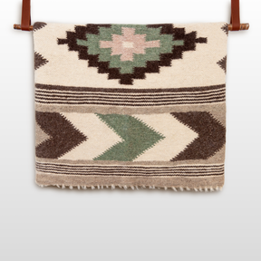 The Myr (Peace) Wool Statement Bed Blanket, featuring geometric patterns in green, brown, and beige, hangs elegantly from a leather strap.