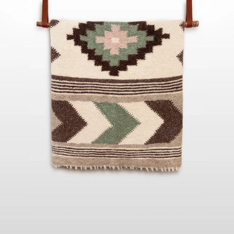 The Myr (Peace) Wool Statement Bed Blanket features handwoven geometric diamond and chevron patterns in green, brown, and beige, displayed hanging on a rod.