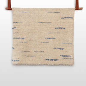 The Horoshy (Kind) Wool Statement Bed Blanket features beige woven wool with scattered blue horizontal stitches, elegantly hanging from two brown leather straps.