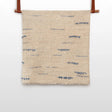 The Horoshy (Kind) Wool Statement Bed Blanket—handcrafted in Ukraine from organic Carpathian sheep wool—features beige textured fabric with sparse blue stitched lines, hanging by brown straps against a white background.