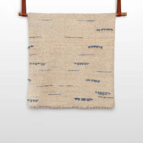 The Horoshy (Kind) Wool Statement Bed Blanket—handcrafted in Ukraine from organic Carpathian sheep wool—features beige textured fabric with sparse blue stitched lines, hanging by brown straps against a white background.