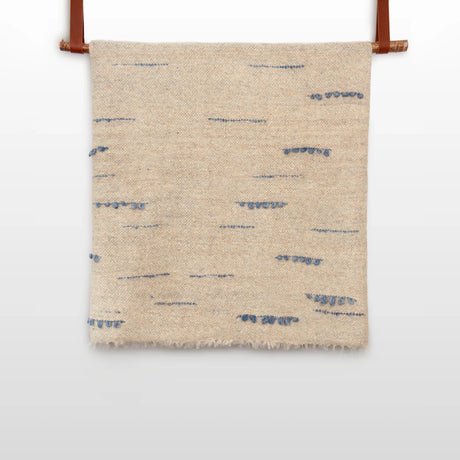 The Horoshy (Kind) Wool Statement Bed Blanket—handcrafted in Ukraine from organic Carpathian sheep wool—features beige textured fabric with sparse blue stitched lines, hanging by brown straps against a white background.