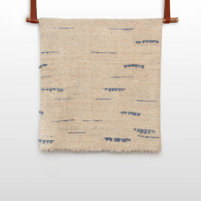 The Horoshy (Kind) Wool Statement Bed Blanket—handcrafted in Ukraine from organic Carpathian sheep wool—features beige textured fabric with sparse blue stitched lines, hanging by brown straps against a white background.