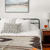 A neatly made bed with white linens, a striped pillow, and the handcrafted Myr (Peace) Wool Bed / Couch Runner featuring a rhombus-dot design beside a wooden nightstand.