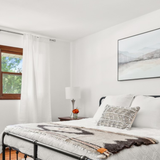 Bright bedroom with white walls, a made bed featuring the Myr (Peace) Wool Bed / Couch Runner, a window with white curtains, and framed landscape art.