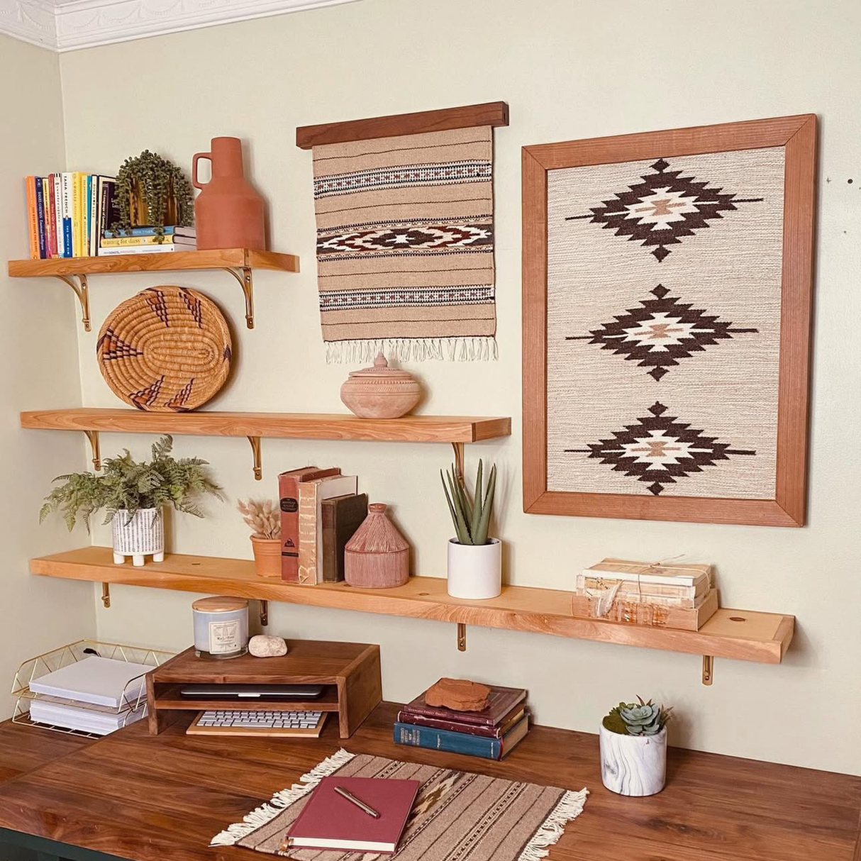Wood shelves display books, pottery, plants, and woven art above a desk styled with books, decor, and the Chitki (Crisp) Wool Rug / Wall Hanging + Wood Hanger ($240 value), adding warmth and eco-friendly mill craftsmanship to the space.