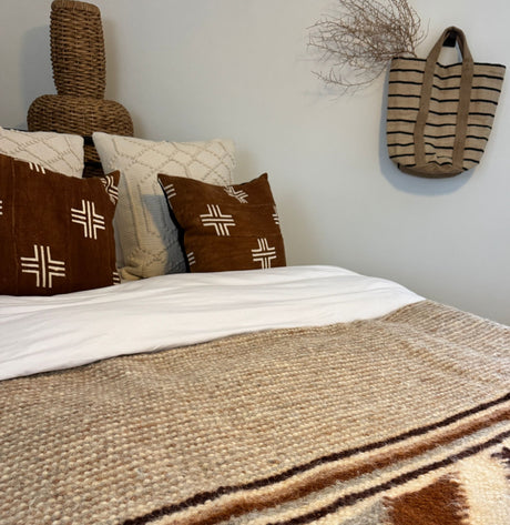 Bed with patterned brown pillows, woven baskets, a striped bag, and the Tkach (Weaver) Wool Statement Bed Blanket against a light wall.