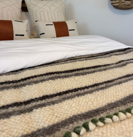 Close-up of a bed with the Faino (Fine) Wool Statement Bed Blanket and pillows, featuring brown and black accents against a white wall.