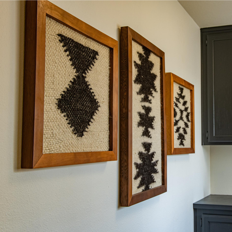 Three pieces of Vira (Faith) Wool Framed Wall Art, each with an abstract black geometric design, are displayed above dark cabinetry against a white wall.