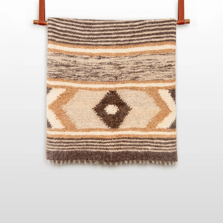 The Peremoha (Victory) Wool Statement Bed Blanket, handwoven with geometric patterns in beige, brown, and tan, hangs from two leather straps against a white background.
