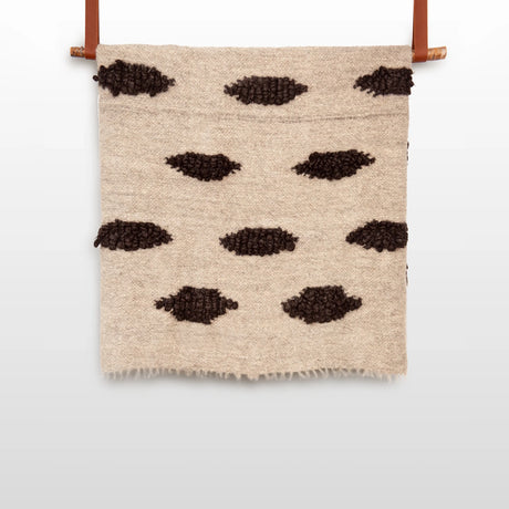 The Khmara (Cloud) Wool Statement Bed Blanket features a woven beige design with rows of dark ovals, inspired by Ukrainian artisan craftsmanship and reminiscent of a handwoven wall hanging.