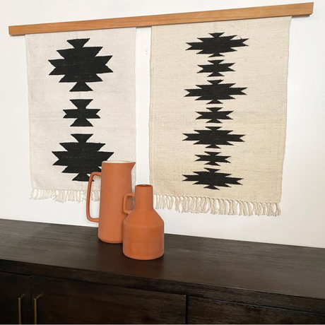 The Chysto (Pure) Wool Rug / Wall Hanging, displayed with a wood rug hanger, features bold black geometric patterns and is part of a striking art ensemble above two terracotta vases on a dark wooden table.