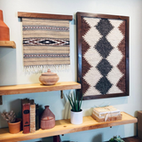 Shelves with pottery, books, plants, and two eco-friendly Dobre (Good) Wool Framed Wall Art pieces featuring geometric patterns on a light wall.