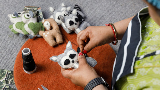 Person sewing a small felt raccoon toy, surrounded by other felt animal toys and sewing supplies.