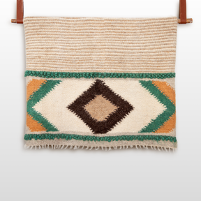 The Tkach (Weaver) Wool Statement Bed Blanket features a cream woven textile with geometric diamond patterns in brown, green, and gold, hanging on a wooden rod—perfect for making a striking statement in any room.