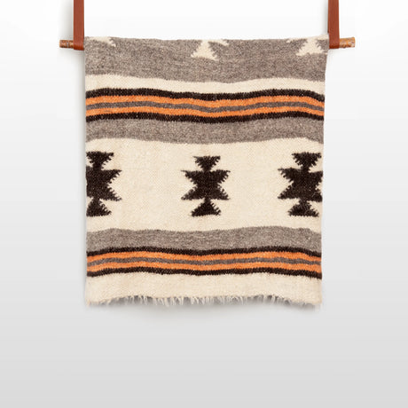 The Chysto (Pure) Wool Statement Bed Blanket is handwoven from organic, cruelty-free Carpathian wool, featuring bold geometric black, brown, and orange stripes and displayed on two leather straps.