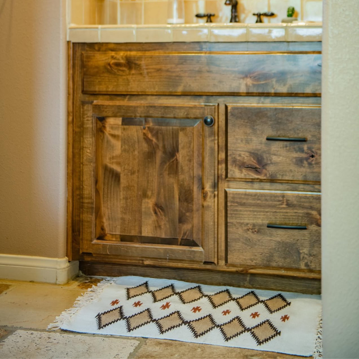The Blazheny (Blissful) Wool Rug with a diamond pattern sits before a wooden bathroom cabinet, bringing a touch of neutral decor to the space.