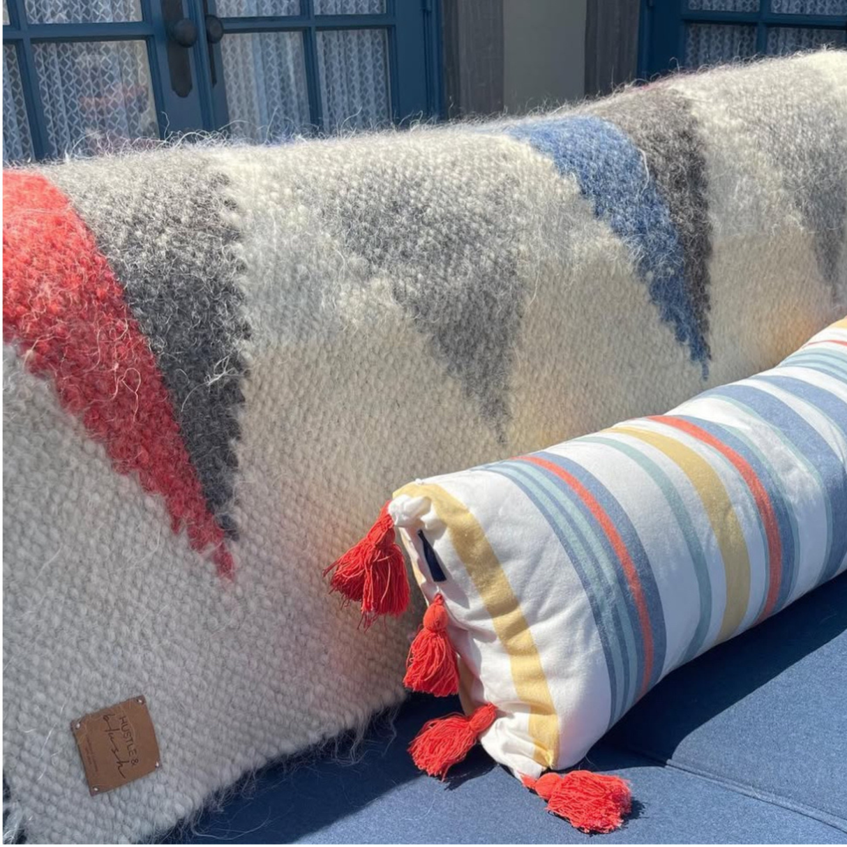 The Harmonia B (Harmony) Wool Statement Throw adds cozy charm to a blue outdoor seat, paired with a fuzzy zigzag pillow and a striped pillow with red tassels.