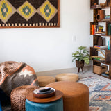 Cozy living room with poufs, throw blanket, bookshelf, plant, and patterned wall art featuring the Mriya (Dream) Wool Wall Hanging for sustainable home decor.