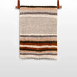 The Mudry (Wise) Wool Statement Baby Blanket, handwoven from organic Carpathian wool with horizontal brown, orange, gray, and beige stripes, hangs on a rod.