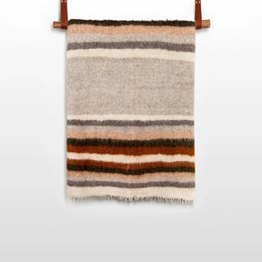 The Mudry (Wise) Wool Statement Baby Blanket, handwoven from organic Carpathian wool with horizontal brown, orange, gray, and beige stripes, hangs on a rod.