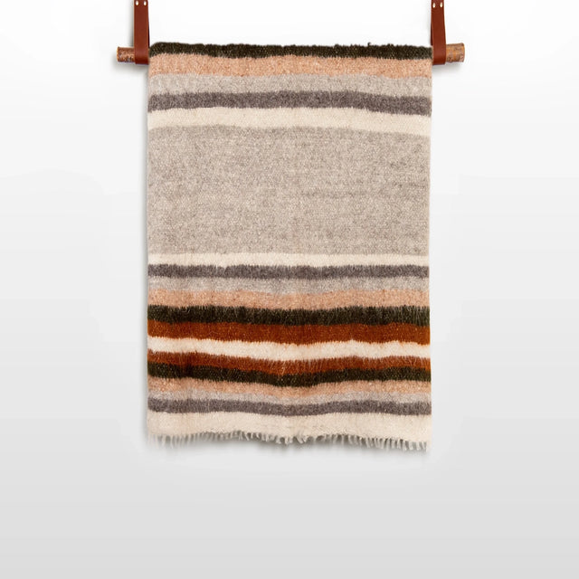 The Mudry (Wise) Wool Statement Baby Blanket, handwoven from organic Carpathian wool with horizontal brown, orange, gray, and beige stripes, hangs on a rod.
