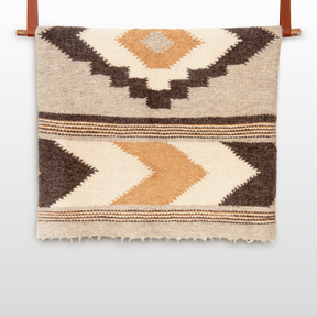 The Myr (Peace) Wool Statement Bed Blanket features handcrafted geometric patterns in brown, tan, and cream, made from undyed yarn and displayed on a leather rod.