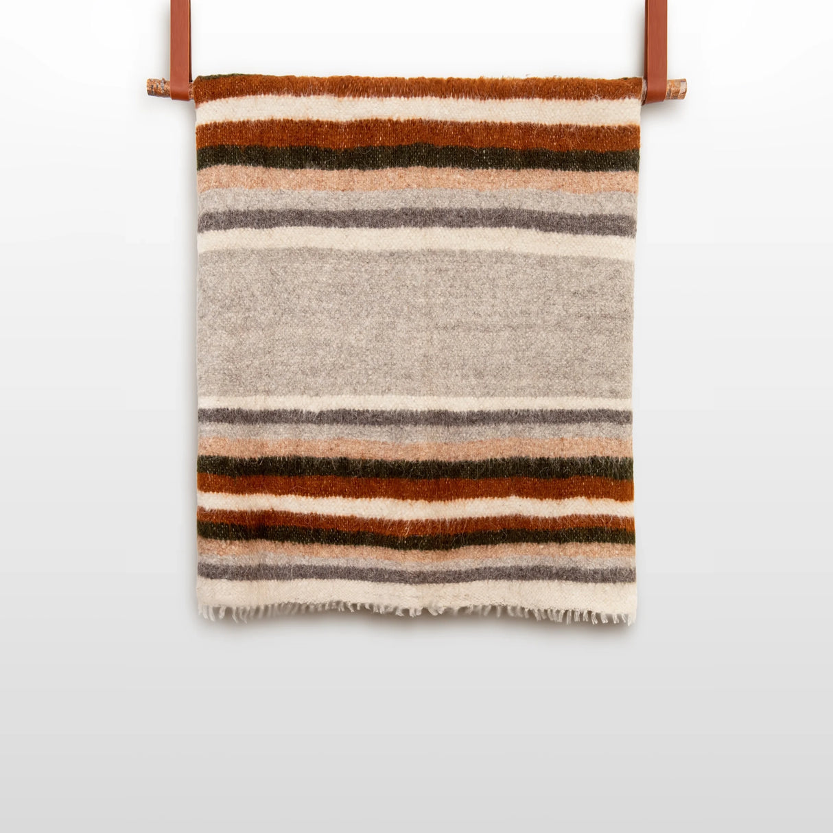 The Mudry (Wise) Wool Statement Bed Blanket, made of organic Carpathian wool and featuring brown, gray, and cream stripes, is folded and hangs on a leather strap against a white wall.
