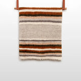 The Mudry (Wise) Wool Statement Bed Blanket, made of organic Carpathian wool and featuring brown, gray, and cream stripes, is folded and hangs on a leather strap against a white wall.